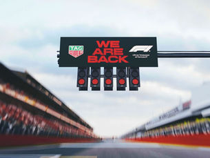 Race track with "WE ARE BACK" sign and traffic lights overhead. TAG Heuer and F1 logos visible. Blurred stands and cloudy sky.