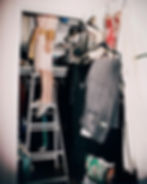 Person in a robe stands on a ladder in a cluttered closet, reaching for an item on an upper shelf. Clothing and shoes are visible.