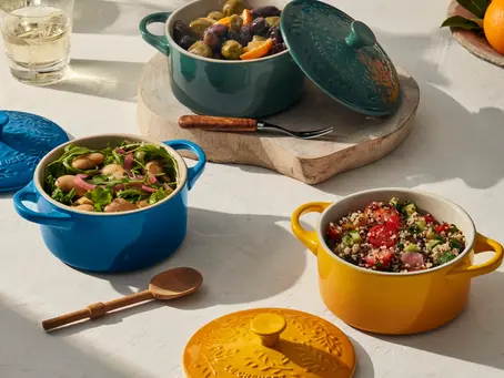 Colorful cookware with food on a white table. Blue, green, and yellow pots with salads and olives. "Le Creuset" embossed lids. Warm sunlight.
