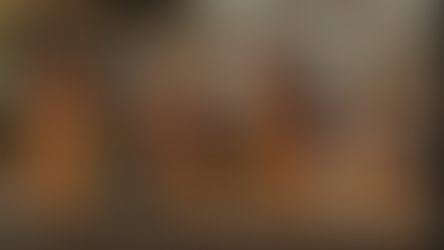 Four women in aprons laugh and hold hands in a kitchen. Shelves with jars in the background. Warm, cheerful atmosphere.