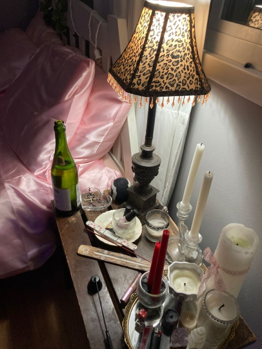 Cozy nightstand with a leopard-print lamp, candles, incense, and a green bottle by pink satin bedding. Warm, intimate ambiance.