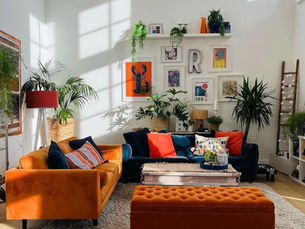 Bright living room with orange sofa, colorful cushions, plants, artwork, and sunlight casting window shadows. Cozy and vibrant ambiance.