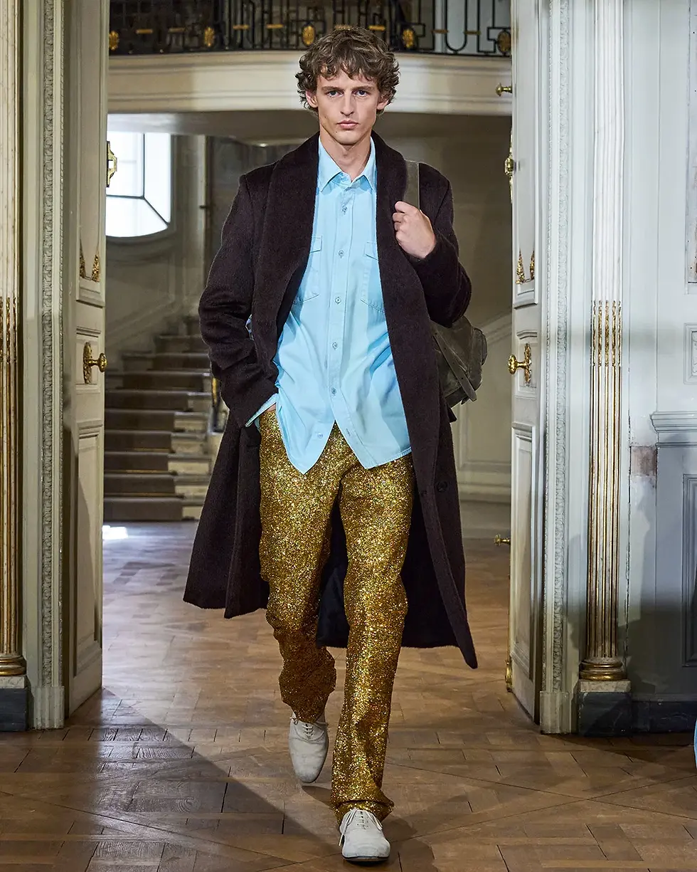 Male model walks through an ornate doorway in a dark coat, blue shirt and gold glitter pants, carrying a backpack on a runway.