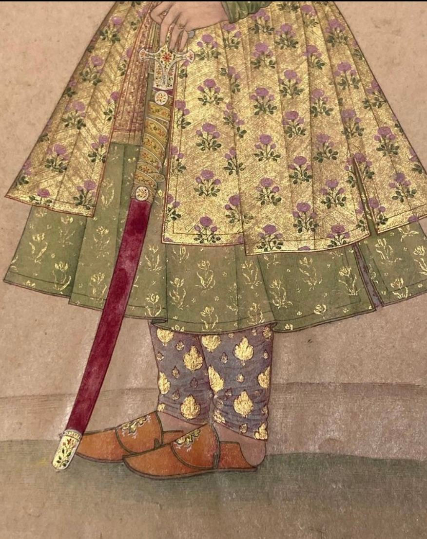 Person in ornate floral attire holds a decorated sword. The robe is gold with purple flowers, and green patterns adorn the lower outfit.