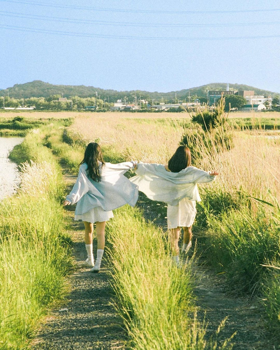Two people in light clothes run joyfully along a grassy path in a sunny rural landscape, with distant hills under a clear blue sky.