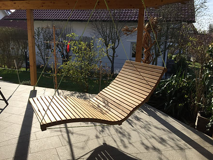 Moderne Holzschaukelliege unter Pergola auf heller Terrasse mit Gartenblick.