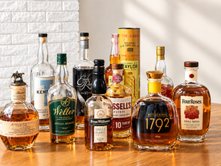 Assorted bourbon bottles, including Blanton’s, Weller, Eagle Rare and Four Roses, arranged on a wooden table against a brick wall.