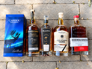Five whiskey bottles and a blue Caribou Crossing box lined up on a sunlit stone patio, with bold labels and warm amber liquor.
