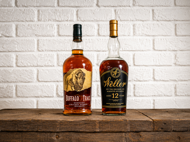 Two classic bourbon bottles, Buffalo Trace and Weller 12 Years, are displayed on a rustic wooden surface against a white brick background, showcasing their rich amber hues and distinctive labels.