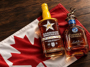 Two whiskey bottles, Garrison Brothers and Caribou Crossing, rest on a Canadian flag on a dark wooden table.