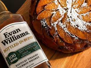 Evan Williams bourbon bottle beside a powdered-sugar-topped Peach Bourbon Upside-Down Cake, on a wooden board, warm rustic close-up.
