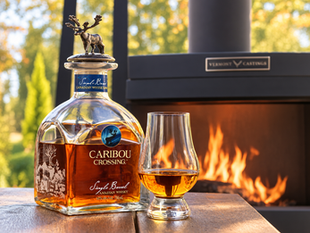 Caribou Crossing single barrel Canadian whisky bottle and glass on a table by an outdoor fireplace, warm sunset glow