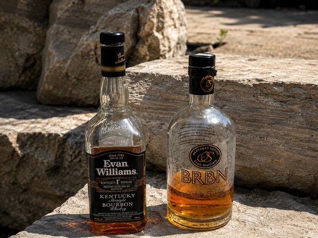 Two whiskey bottles, one labeled "Evan Williams Kentucky Straight Bourbon" and the other "Okanagan Spirits BRBN Family Reserve," sit side by side on a sunlit stone surface.