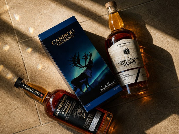 A beautifully composed image showcases a trio of distinctive whiskey bottles, highlighting the rich amber tones of Caribou Crossing, Barnburner Whiskey, and John Sleeman & Sons Spring Mill Distillery. The arrangement captures the play of light and shadow, emphasizing the allure and craftsmanship of each unique label.