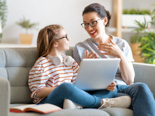 a child sat on a sofa with a laptop on her knee, talking to her mother