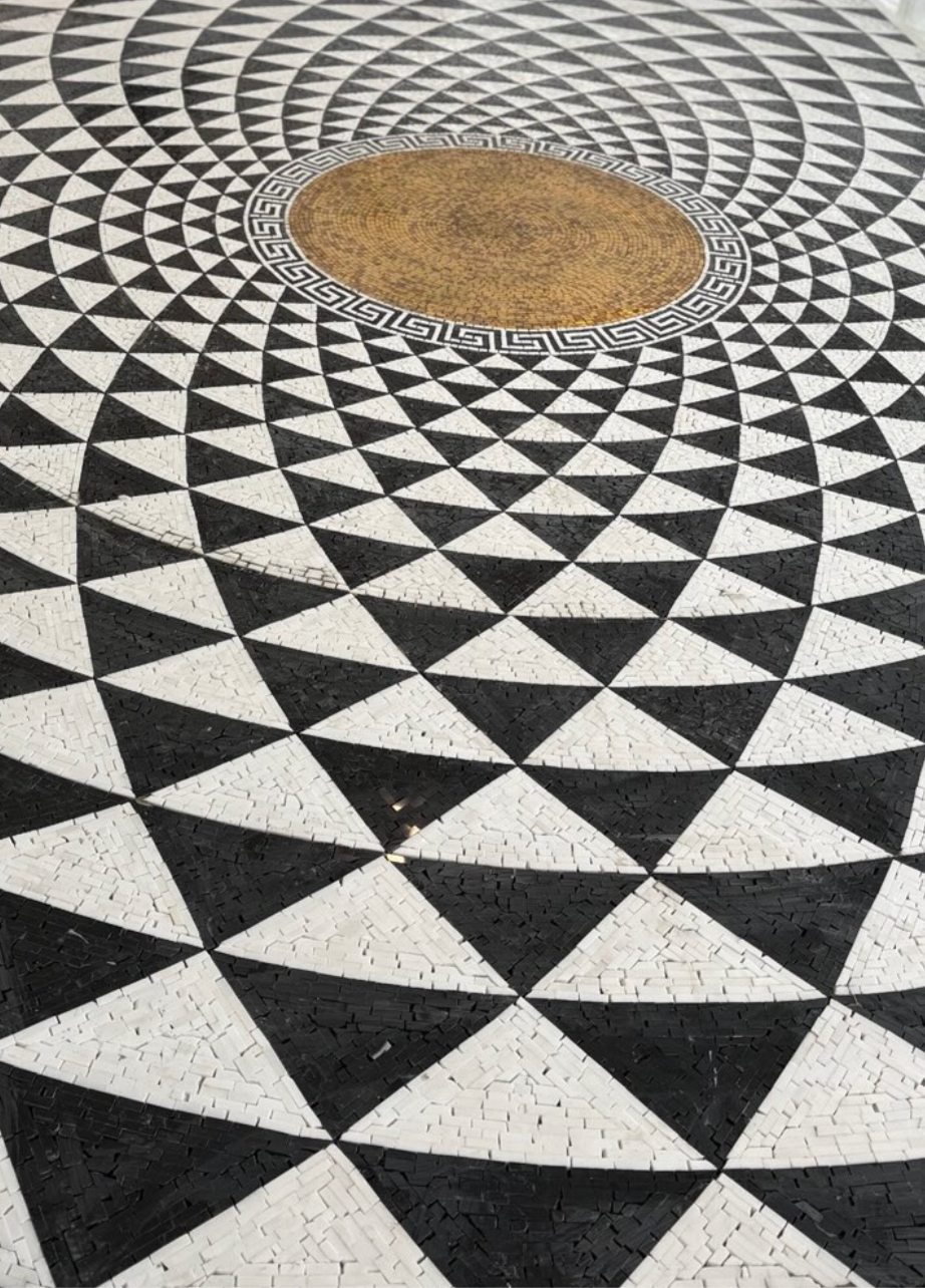 Pavimentazione musiva artistica per interni residenziali.
Sviluppo Visivo: Un disegno optical geometrico a moduli triangolari che si sviluppa in cerchi concentrici attorno a un nucleo centrale perfetto. Il perimetro del medaglione è nobilitato da un classico motivo a greca, che fa da ponte tra l'estetica contemporanea e la storicità della tradizione musiva.
