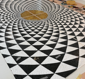 Pavimentazione musiva artistica per interni residenziali. Un disegno optical geometrico a moduli triangolari che si sviluppa in cerchi concentrici attorno a un nucleo centrale perfetto. Il perimetro del medaglione è nobilitato da un classico motivo a greca, che fa da ponte tra l'estetica contemporanea e la storicità della tradizione musiva.