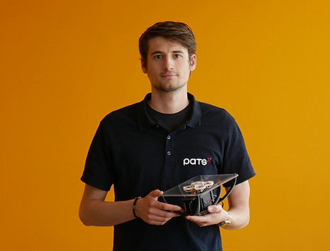 Man wearing a PATS shirt holding a small PATS-X drone prototype against an orange background