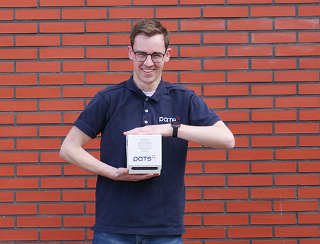 Man wearing a PATS shirt holding a PATS-C device with both hands in front of a brick wall