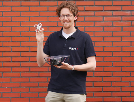 Man wearing a PATS shirt holding a small drone in one hand and a larger drone frame in the other, standing in front of a brick wall