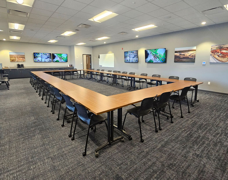 Meeting room with grey carpet, brown rolling desks, black rolling chairs and multiple tvs on all 4 white walls.