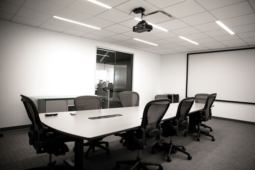 Large white table with 10 rolling chairs surrounding it. The table is on grey carpet and sits in front of a white wall with a projector screen.