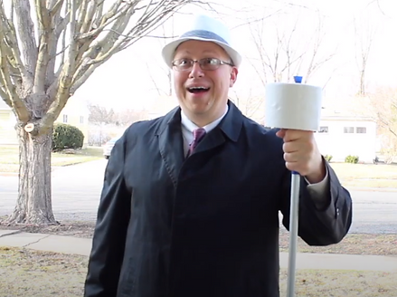 Poster image for H.O.M.E. Invasion.  A man grinning with an open mouth.  He is wearing a fedora and suit.  He is holding a pole with a roll of toilet paper stacked on the end.