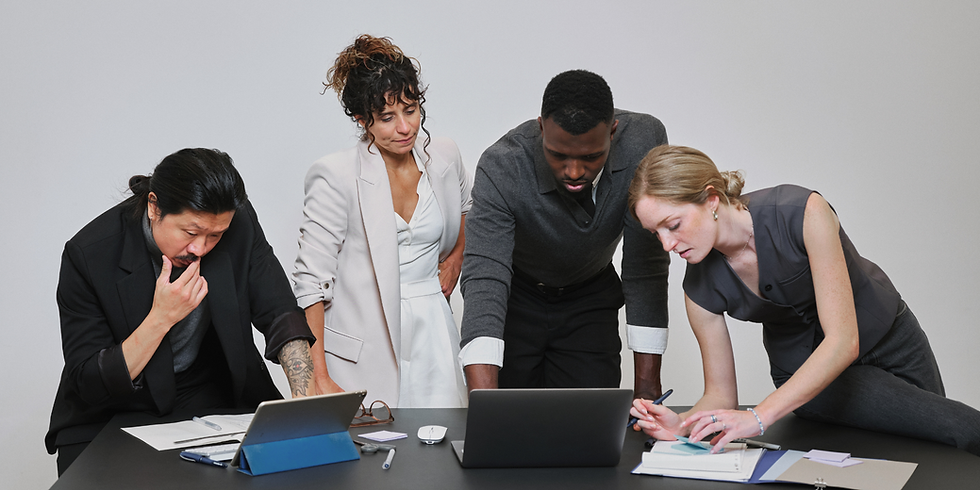 A diverse creative team leaning over a black desk, intensely collaborating on a project with a laptop, tablet, and handwritten notes against a plain grey background.