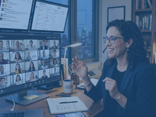 A female remote leader wearing a headset smiles and gestures during a large video call at a multi-monitor desk setup.