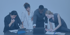 A diverse creative team leaning over a black desk, intensely collaborating on a project with a laptop, tablet, and handwritten notes against a plain grey background.