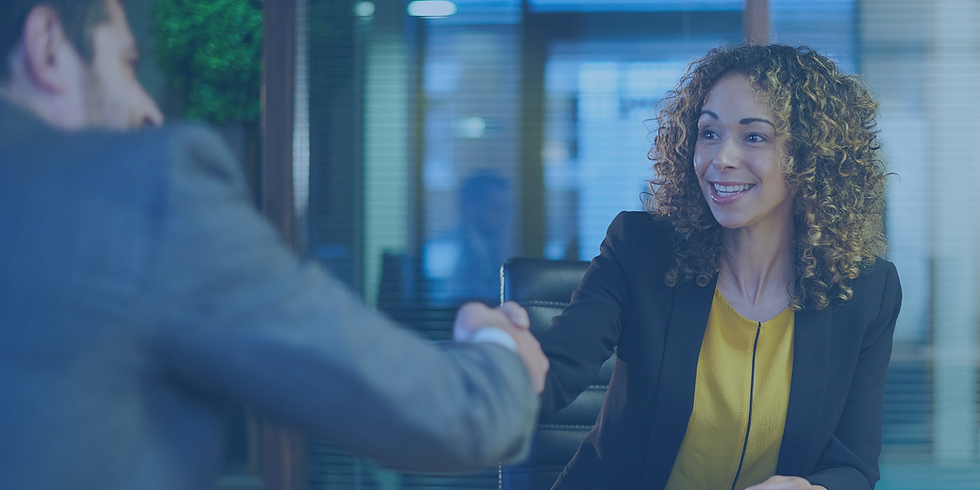 A smiling woman in a black blazer and yellow top shakes hands with a colleague in a modern office.