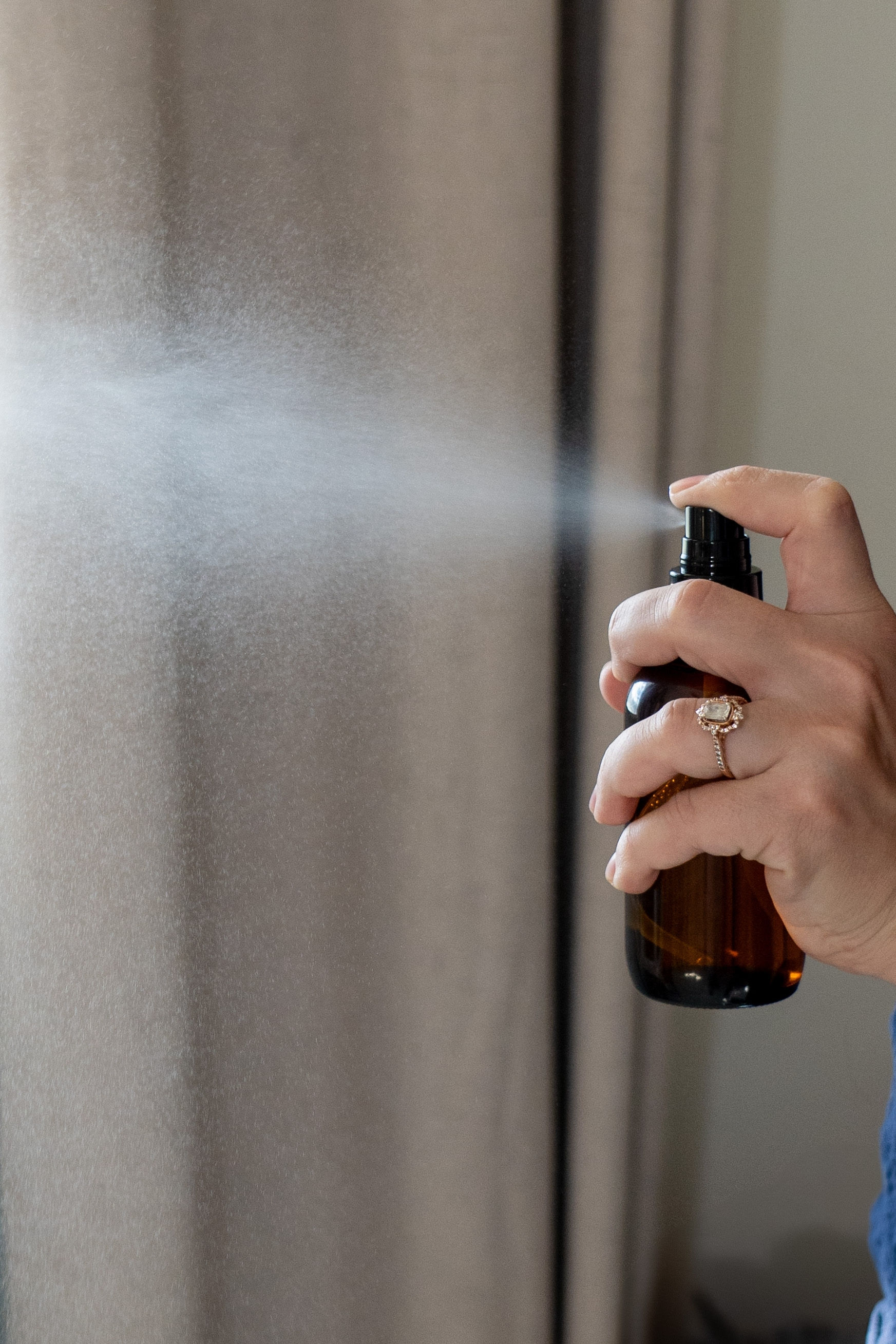 Person spraying a brown bottle, mist in front of curtains and a wall.