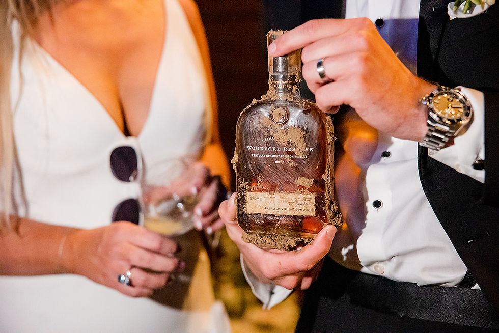 A person in a suit holds a mud-covered Woodford Reserve whiskey bottle. A woman in a white dress holds a glass. Both appear celebratory.