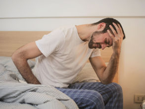 Man sitting in bed holding his head in discomfort, appearing stressed or in pain.