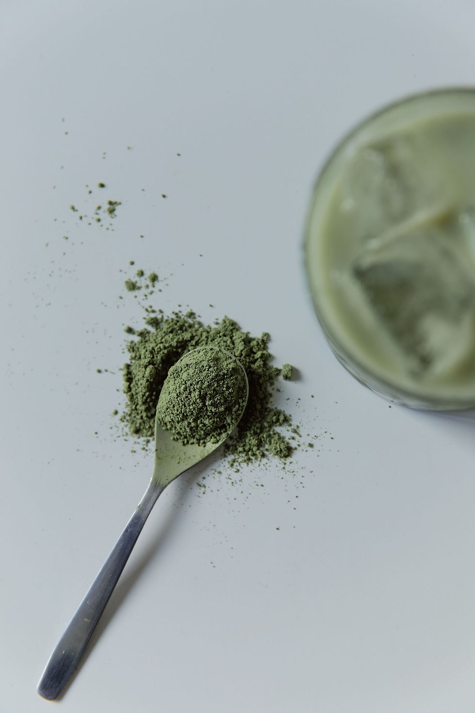 A close-up of a spoonful of weird powder (green) on a metal teaspoon, next to a green drink which may or may not be a smoothie.