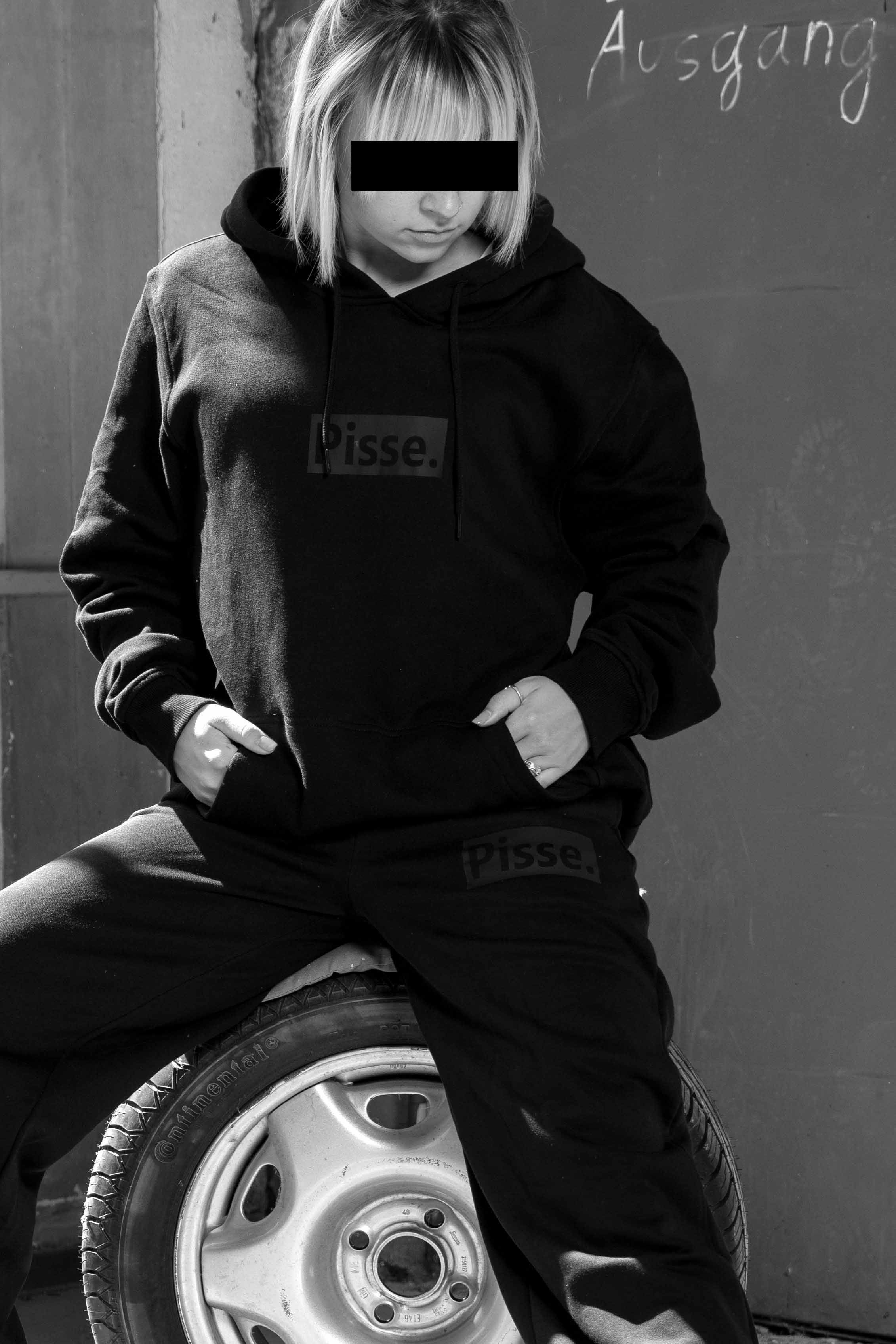 Frau mit schwarzem Hoody. Sie guckt zur Seite. Auf dem Hoody is "Pisse." zu lesen.