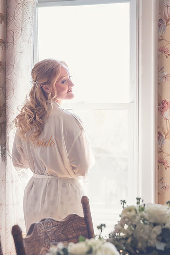 Bride in a white robe stands by a window, side glance, with flowers visible. Soft lighting and floral curtain create a serene mood at a Maine Wedding