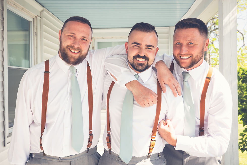 Groom and Groomsmen in white shirts, mint ties, and suspenders smile on a porch. Light background with trees, conveying a joyful mood at Maine Wedding.
