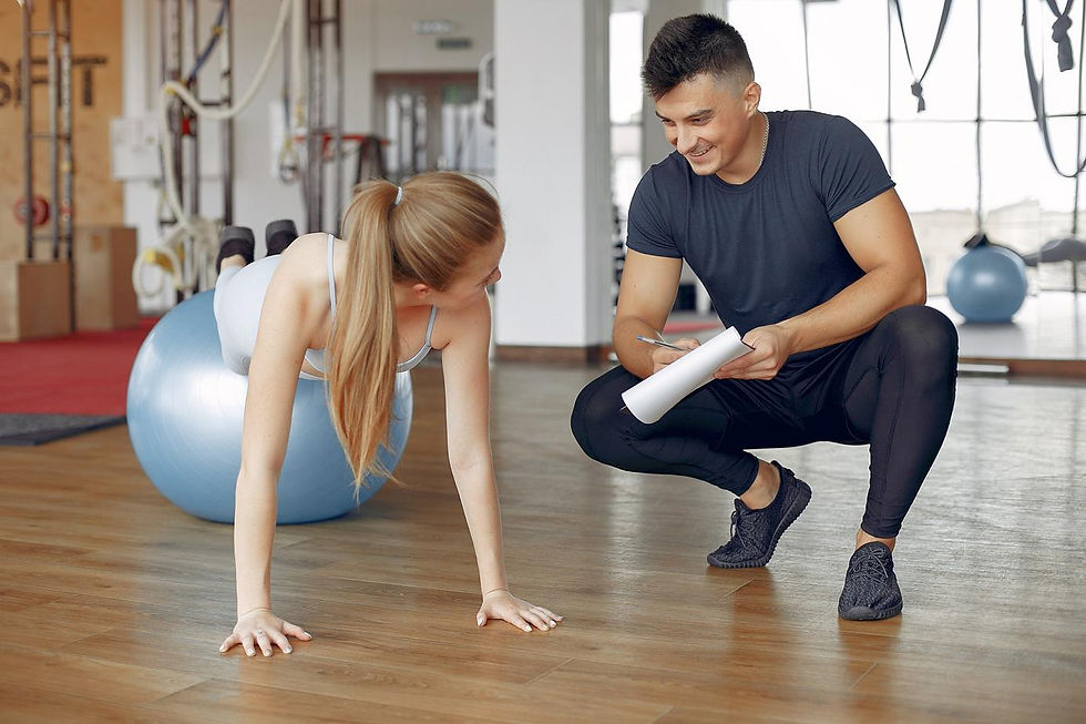Un coach sportif est accroupi, un carnet de notes à la main. Il donne des conseils à une jeune femme qui fait des pompes, les pieds surélevés sur un swissball
