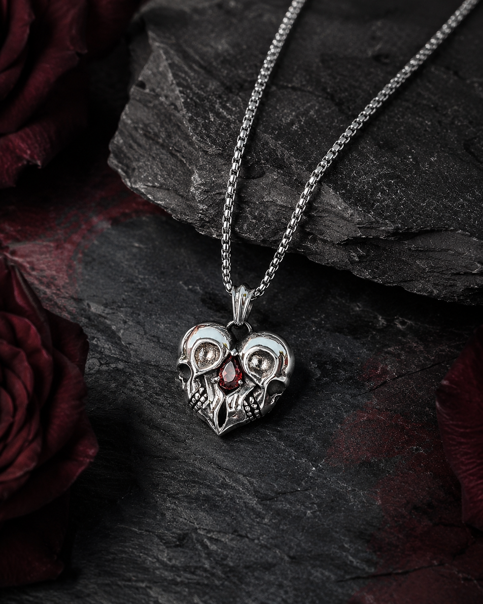 Thumbnail: Twin skull heart stainless steel necklace with red teardrop stone displayed on dark stone with red roses.