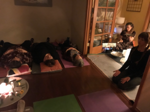 Several participants lie on yoga mats in a dimly lit room at Hoodland Bazaar’s Phoenix Room during a sound bath, while a Reiki practitioner leads the session surrounded by bowls and candles.
