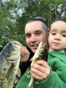 A father and his young son smile and show off two freshly caught bass, celebrating a successful double catch during a hot bite in the Pacific Northwest.