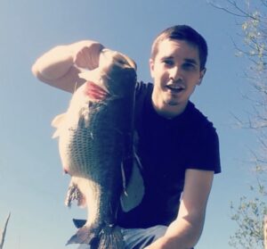 A smiling angler displays a massive Largemouth Bass against a clear blue sky—his personal best catch in Washington state waters.