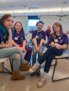 Four 5th grade students from Welches School sit together in matching purple Oregon Battle of the Books shirts, smiling confidently as they wait to compete at the OBOB 2025 state tournament.