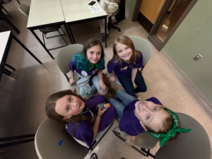 A top-down view of the Welches Reading Rizzlers team huddled together in a circle of chairs, grinning up at the camera before their Oregon Battle of the Books match.