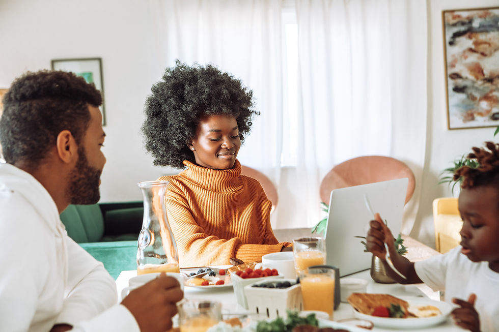 A mother, father, and their child sit at the breakfast table. In front of them is a spread of fruits, orange juice, and pancakes. While the child and father eat, the mother sits at the head of the table with a smile on her face as she works on her laptop.