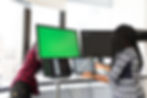 A Black woman stands with two open laptops and a dual monitor system at a standing desk, while another person stands behind one of the dual monitors, appearing to be plugging something in. They are both in a brightly-lit office space with large windows.
