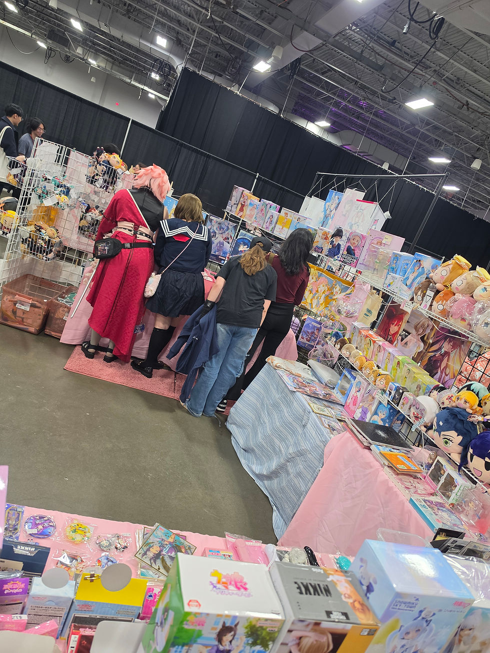 Wide-angle view of a cozy anime convention aisle with a colorful photo table and fans browsing nearby.