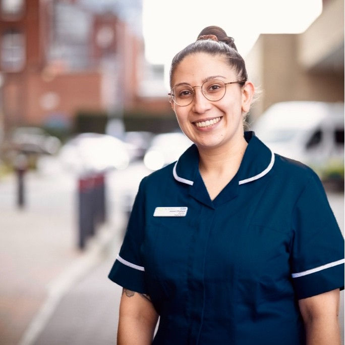 Image of Jessie Dhaliwal wearing her nurses uniform and glasses outside a hospitalwith the background blurred
