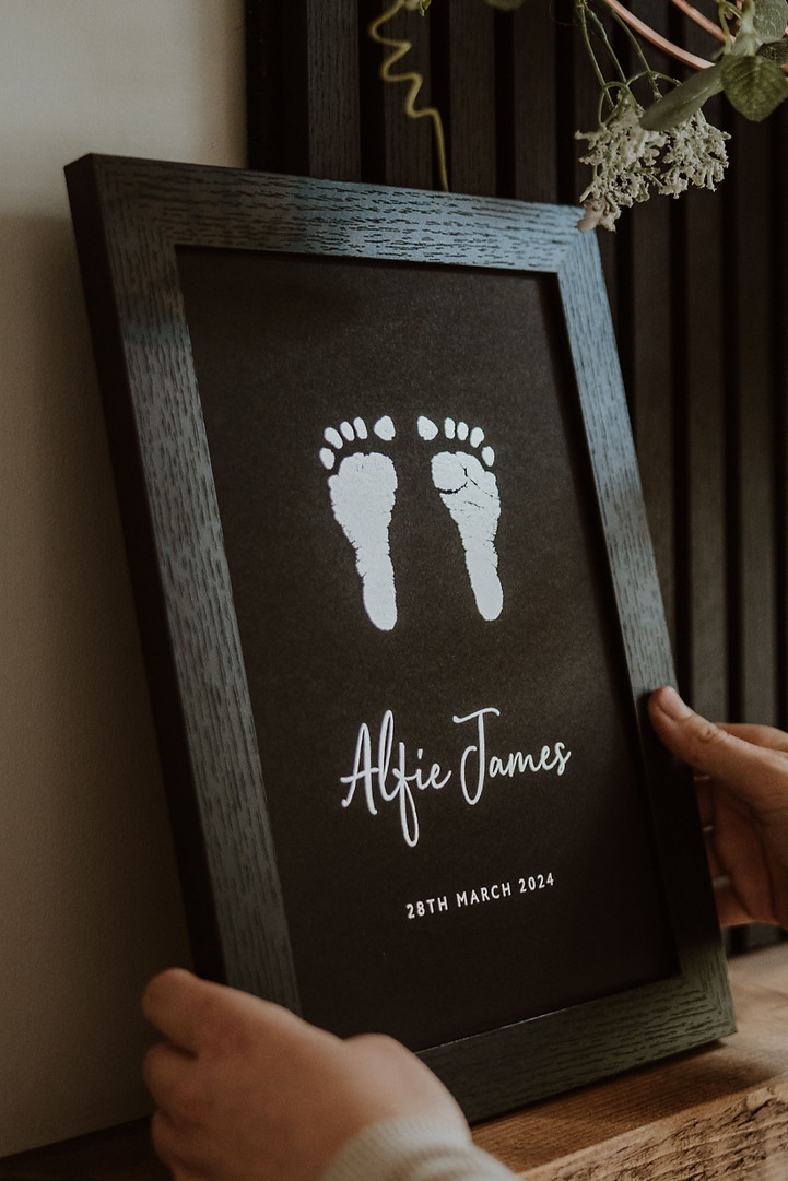 Hands holding black framed silver foil print with baby footprints